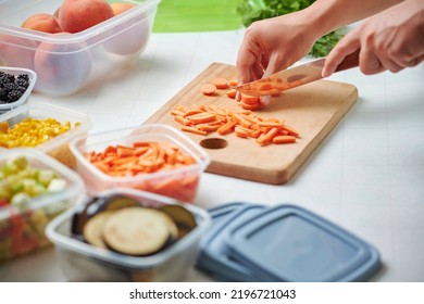 Hands Of Young Female Chopping Fresh Carrot On Wooden Board While Preparing Vegetables For Freezing. Plastic Containers With Raw Cut Vegetables For Freezing On Kitchen Table