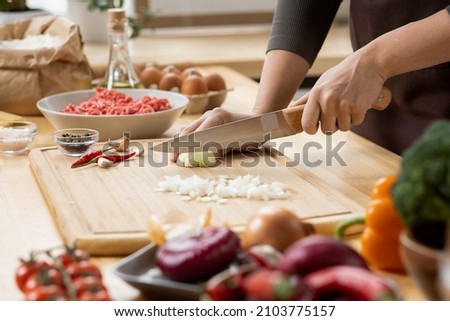Hands of young female chopping fresh onion on wooden board while preparing italian pasta with vegetables and minced meat