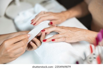 Hands of young girl manicurist perform trimming saws nails painted with red varnish to elderly lady client. Close-up, high angle of view