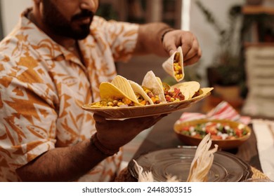 Hands of young Hispanic man holding plate with tasty Mexican tacos and taking one of them while sitting by served festive table at dinner - Powered by Shutterstock