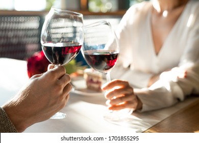 Hands of young man and his girlfriend clinking with wineglasses over table while enjoying romantic date and lunch in luxurious restaurant
