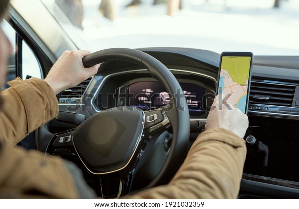 Hands of young man in warm winterwear holding by\
steer while sitting in car and pointing at map on screen of\
smartphone during travel