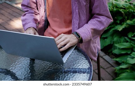 the hands of a young programmer working on a silver laptop, sitting at a glass table in a summer cafe in the park. The concept of remote work