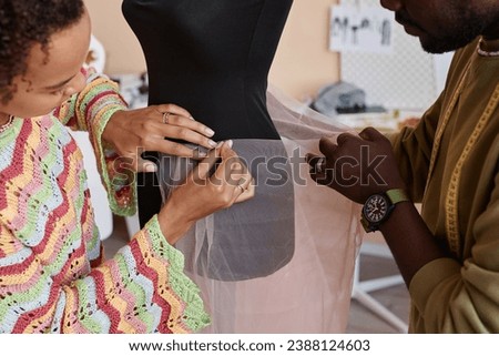 Hands of young seamstress pinning piece of pink fatin fabric to dummy while male colleague helping her during work over new collection
