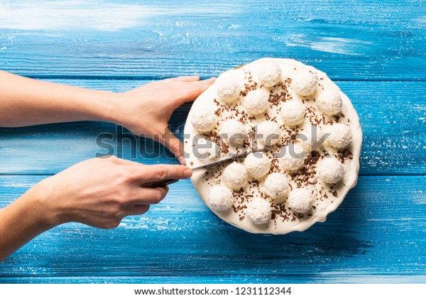 The hands of a young woman cut the cake into pieces.\
Celebratory cake with white cream cream decorated with round\
candies with coconu chips on a blue background. Copy space. View\
from above. 