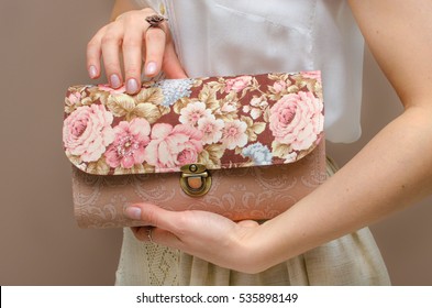 In The Hands Of A Young Woman Dressed In White Cotton Blouse And Beige Linen Skirt, A Delicate Clutch Bag With Floral Print Of Roses On A Brown Background. Women's Day, Fashion, Spring, Romance, Love.