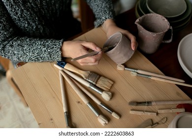 Hands of young woman master of ceramics paint pottery cup or bowl on a background of art studio. Brushes are scattered on the wooden table. Top view - Powered by Shutterstock