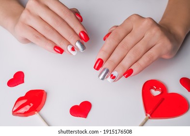 The Hands Of A Young Woman. The Nails Are Covered With Red Gel Polish For Valentine's Day. On A White Background With Lollipops In The Shape Of A Heart And Lips. Nail Art And Design Ideas.