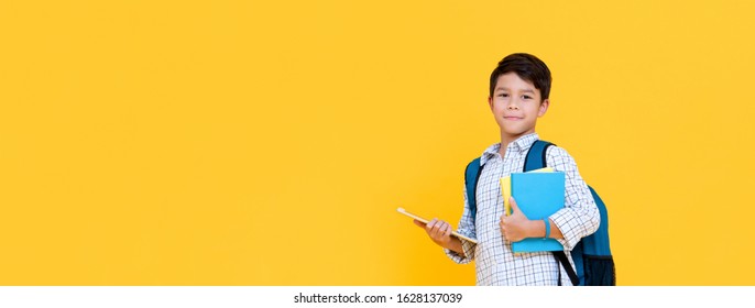 Handsome 10 year-old boy with backpack holding books and tablet computer in yellow banner background with copy space for education concept