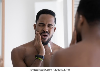 Handsome 30s African Guy With Frown Face Expression Reflected In Mirror Touch Cheek While Brush Teeth, Feeling Pain Due Oral Dental Problems. Toothache, Sensitive Enamel Need Repair Toothpaste Concept