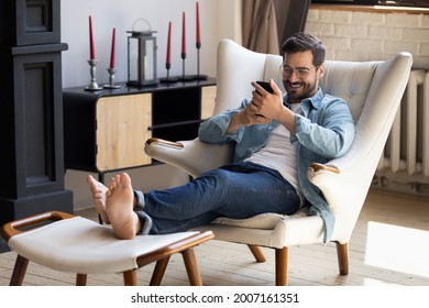 Handsome 30s single man relaxing on comfy armchair put feet on footstool holding in hands smartphone spend weekend alone in cozy cottage house. No stress, home owner, modern tech usage, fun concept