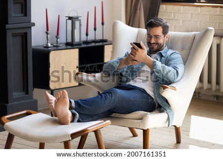 Handsome 30s single man relaxing on comfy armchair put feet on footstool holding in hands smartphone spend weekend alone in cozy cottage house. No stress, home owner, modern tech usage, fun concept