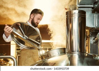 Handsome, adult brewer inspecting process of brewing beer with steam. Confident, bearded specialist opening cover of metallic brew kettle, looking down.