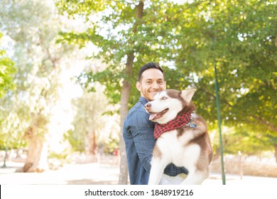 Handsome Adult Man Carrying In His Arms A Big Brown Dog And Surrounded By Green Trees At The Park