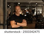 Handsome adult personal trainer or fitness coach standing wearing black t-shirt with cross hand at gym. Motivation for regular workout, body building, health and energy. Crafting my workout routine.