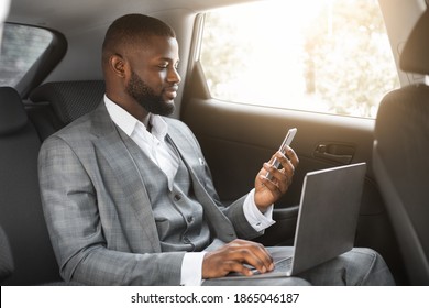 Handsome African American Businessman With Laptop Holding Smartphone While Going By Car On Business Trip, Copy Space. Black Entrepreneur Looking At Mobile Phone Screen, Using Laptop