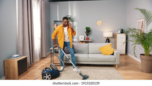 Handsome African American Cheerful Young Guy Vacuuming Living Room And Smiling Speaking On Smartphone, Male Working Cleaning House Using Vacuum While Calling On Cellphone, Housekeeper Concept