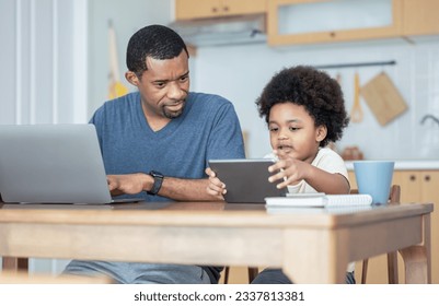Handsome African American father using laptop computer and his cute little boy using digital tablet learning online, watching video while sitting on table in kitchen room at home. Technology concept. - Powered by Shutterstock