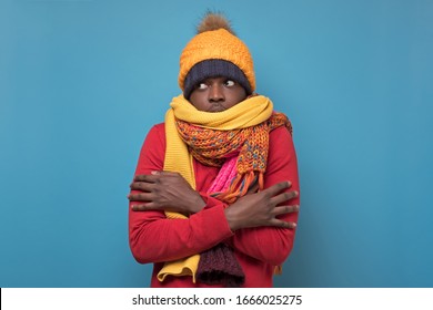 Handsome african american funny man in several hats and scarfs on blue background. Winter fashion.