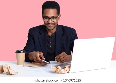 Handsome African American Male Sits At Desk With Lap Top And Crumpled Shirts Of Paper, Works Online. Dark Skinned Guy Takes Smart Phone And Looks Down On It, Drinks Coffee Or Tea. Technology Concept.