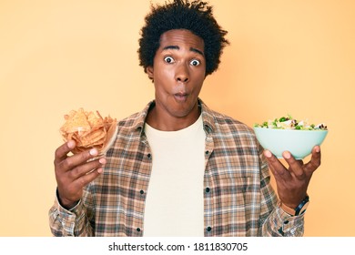 Handsome African American Man With Afro Hair Holding Nachos And Healthy Salad Making Fish Face With Mouth And Squinting Eyes, Crazy And Comical. 