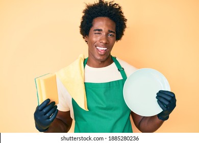 Handsome African American Man With Afro Hair Wearing Apron Holding Scourer Washing Dishes Winking Looking At The Camera With Sexy Expression, Cheerful And Happy Face. 