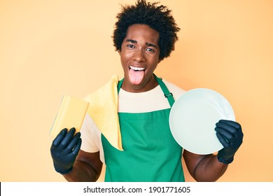 Handsome African American Man With Afro Hair Wearing Apron Holding Scourer Washing Dishes Sticking Tongue Out Happy With Funny Expression. 