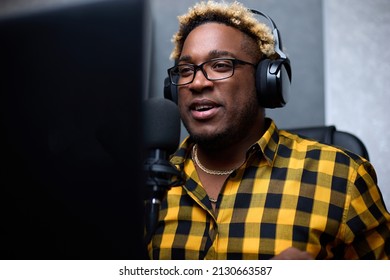 Handsome African American Man Broadcasts His Audio Podcast In Small And Cozy Home Broadcasting Studio. Smiling Black Man In Glasses And A Plaid Shirt Is Broadcasting Using A Microphone And Headphones