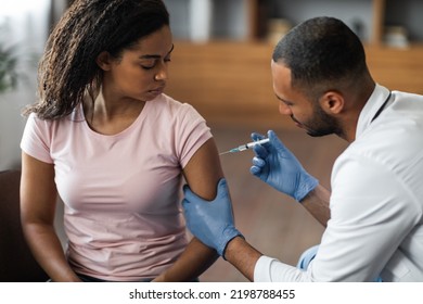 Handsome African American Man Making Injection In Shoulder For Female Patient, Wearing Blue Gloves. Young Black Lady Getting Vaccinated Against Coronavirus At Clinic, Closeup Shot