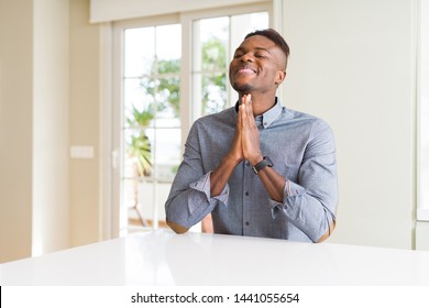 Handsome african american man on white table praying with hands together asking for forgiveness smiling confident.