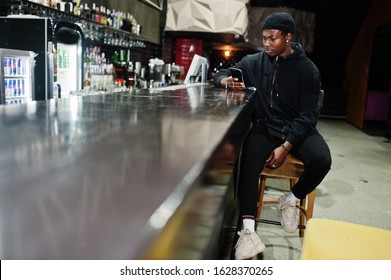 Handsome African American Man Posing  Inside Night Club In Black Hat, Sitting On Bar Counter.