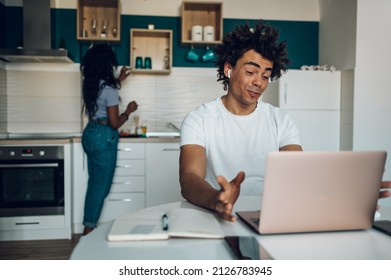 Handsome african american man sitting in the kitchen and making video call with his colleagues while using laptop and ipods. Remote working from home with technology.