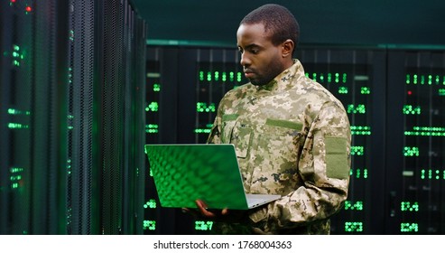 Handsome African American Man From US Army Typing On Keyboard Of Laptop Computer While Checking Data At Servers Monitors In Dark Room. Male Technician Of Anti-terrorism Center.