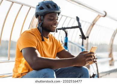 Handsome african american man wearing safety helmet, holding mobile phone, online shopping outdoors sitting near bicycle. Young student wearing stylish casual orange t shirt checking mail - Powered by Shutterstock