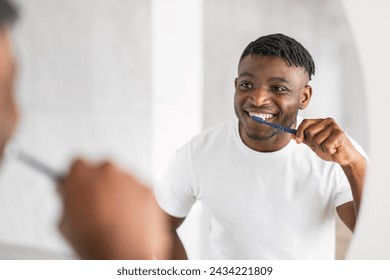 Handsome African American millennial guy brushing teeth with toothbrush stands by the mirror, caring for his oral health in modern bathroom. Daily dental care routine. Selective focus - Powered by Shutterstock