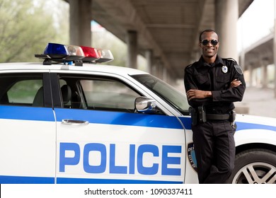 handsome african american police officer with crossed arms leaning back on car and looking at camera