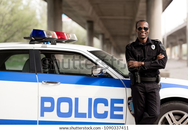 handsome african american\
police officer with crossed arms leaning back on car and looking at\
camera