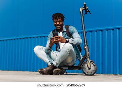 handsome African American sitting on an electric scooter, with a phone in his hands, and typing an SMS message, 5g internet, online correspondence, against background of a blue building