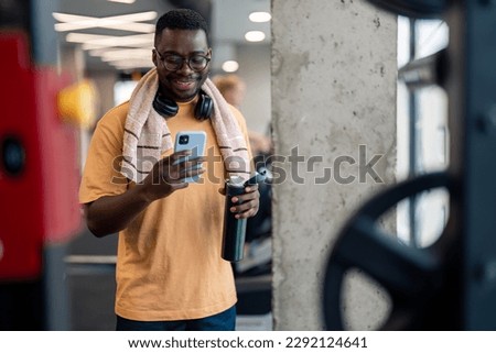 Handsome African American sportsman using mobile phone while taking a break from exercising, standing in gym and smiling. Young fit male fitness instructor texting on mobile phone while resting in gym