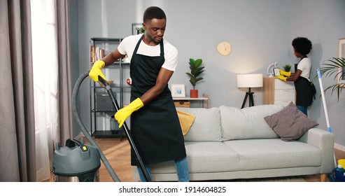 Handsome African American Young Man In Black Apron Working Vacuuming Floor In Room, Female Worker Wiping Dust From Furniture Behind, Cleaning Service, Professional Cleaning, Job Concept