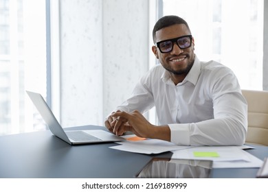 Handsome African Businessman Sit At Desk With Laptop Smile Staring Aside In Skyscraper Office Boardroom. Successful Employee, Career Growth, Modern Tech, Business Owner Portrait, Aspirations Concept