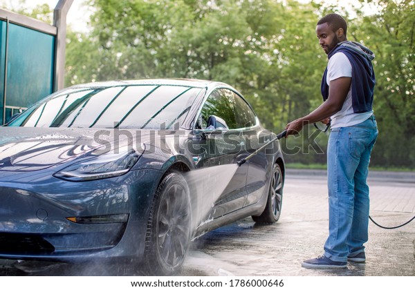 Handsome African guy in t-shirt and jeans
washing his blue modern electric luxury car, rinsing the soap with
high pressure water jet. Manual car wash with pressurized water in
car wash outside.
