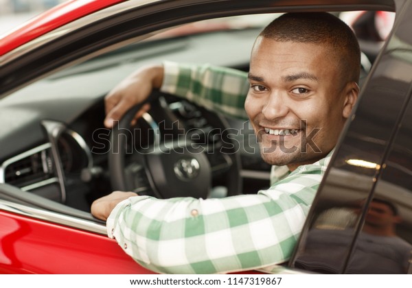 Handsome African male driver smiling to the\
camera, while sitting in his new car. Happy man enjoying driving\
his new automobile. Male customer sitting in a modern auto at the\
dealership. Sales\
concept