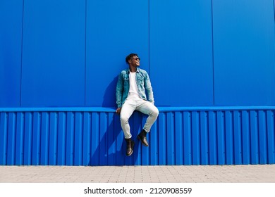 Handsome African Male Fashion Model In Sunglasses Smiling And Posing While Standing Near Blue Wall Outdoors. Stylish Curly African Man Smiling Over Isolated Blue Background