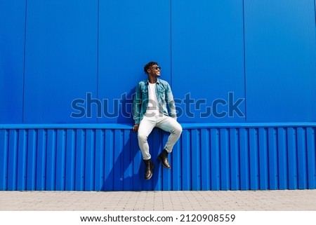 Handsome african male fashion model in sunglasses smiling and posing while standing near blue wall outdoors. Stylish curly african man smiling over isolated blue background