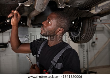 handsome african male repairing bottom of car, check and examine all details. hardworking man in uniform at work