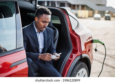 Handsome african man in business suit with money cash in hands sitting inside electric car that is charging. Concept of people, transport and savings.