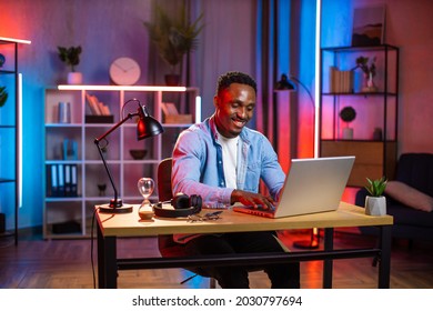 Handsome african man in casual clothes sitting at table and typing on wireless laptop. Young guy doing remote work on portable computer during evening time at home. Freelance concept.