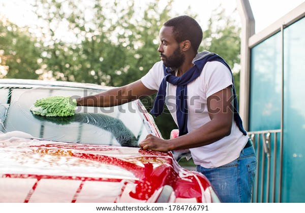Handsome African\
man cleaning his red car windshield with green sponge and soap foam\
outdoors at car wash\
service