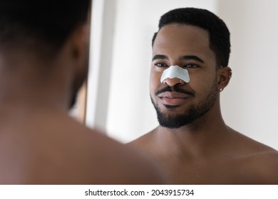 Handsome African man portrait with pore cleansing, anti-blackhead patch strip on nose looking in mirror. Skincare, hygiene, deep cleaning modern effective procedures, beauty treatment routine concept